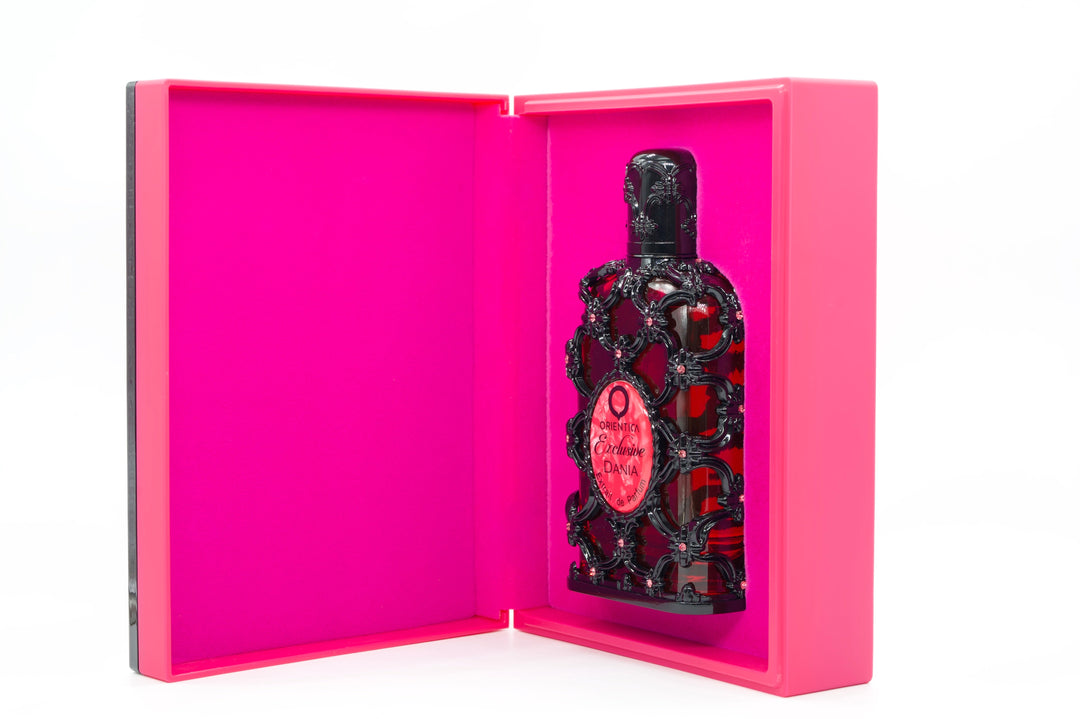 A dark, ornate bottle with a red circular label displayed inside an open hot pink presentation box on a white background.
